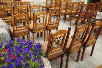 Wooden chairs with patterns on the backs stand in rows on paving stones.