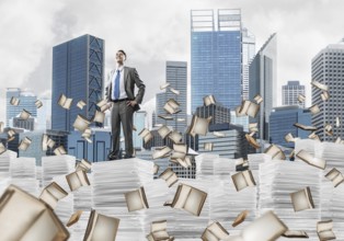 Confident businessman in suit standing on pile of documents among flying books with cityscape on