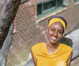 Smiling woman in a yellow dress with a headband, standing near a tree and brick wall, Taken in