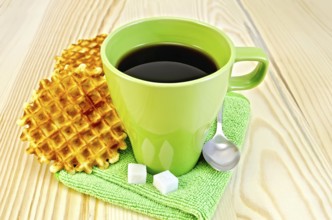 Two golden round waffles, green mug, sugar, spoon on green napkin on a background of wooden board