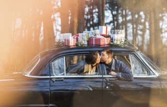 Loving man and woman sticking out of retro car windows and kissing with Christmas gifts on roof in