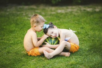 Two children, Caucasian brother and sister, sitting on green grass in backyard of house and hugging