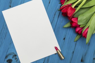 Red tulips, lipstick and a white sheet of paper for your greetings on the background of blue wooden
