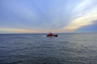 An orange-coloured pilot boat sails on the calm sea at sunrise under a blue sky, 27472 Cuxhaven,