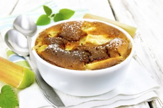 Pie clafouti with rhubarb in the bowl, mint and rhubarb stems on a napkin on wooden plank