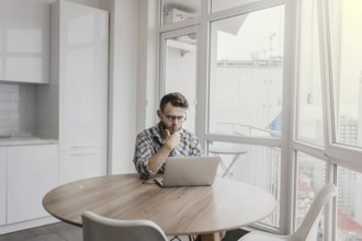 Man in a checkered shirt thoughtfully working on a laptop in a bright workspace