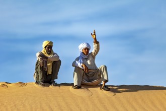 Two men sit on a sand hill in the desert under a clear blue sky, two Tuareg sit on a sand dune and