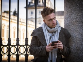 Handsome trendy man wearing dark coat standing and looking down at a cell phone that he is holding,
