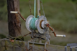 A close-up of an old, rusted winch with green rope attached, mounted on wooden pillars, the