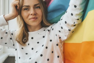 Young blonde millennial hippie woman with rainbow LGBTQ flag at home balcony. Peace and freedom the
