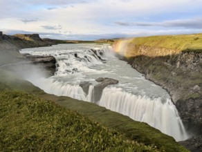 Majestic waterfall cascading over cliffs with rainbow and lush greenery under a cloudy sky. Iceland