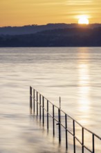 Rising sun over calm lake, railing extends into the water, lido, Litzelstetten, Lake Constance,