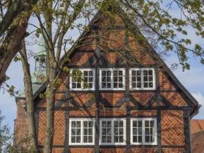 The picture shows a half-timbered house with red bricks, surrounded by trees. A church spire is