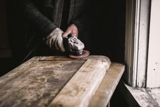 Close-up of hands sanding a wooden door in a dimly lit workshop next to a window