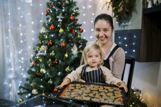 Smiling mother with little daughter sitting at the table with a baking sheet of raw Christmas