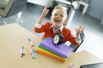 Cute blonde caucasian kid sit at the table with colorful folders, hold alarm clock, evil laugh,