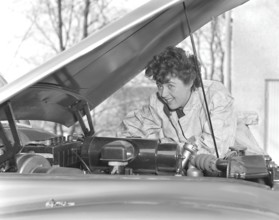 In the 1950s. A woman in an overall fixing something on the car that has it's hood open. Sweden