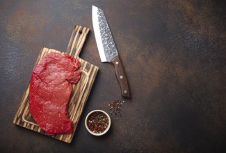 Raw uncooked top round beef steak on wooden cutting board with big kitchen knife and pepper on dark