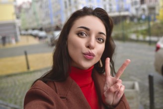 Charming smiling lady in a brown autumn coat takes a selfie on the street with a peace sign
