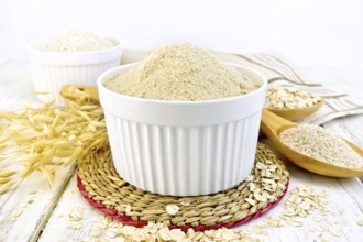Oat flour in white bowl, oatmeal and bran in a spoon, stalks of oats, napkin against the background