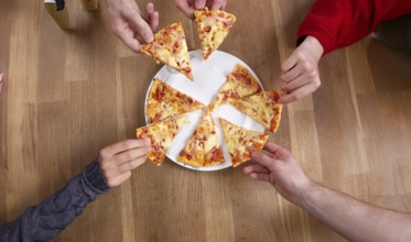 Multiple hands taking slices from a pizza placed on a wooden table