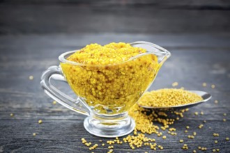 Mustard Dijon sauce in a glass sauceboat and mustard seeds in a spoon on a black wooden board