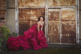 Woman in cloudy red dress is posing over rust wall. Fashion portrait without retouch, only make up