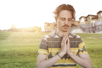 Young man meditating outdoors in the park, sitting with eyes closed and his hands together. man