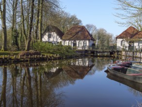 Painting of quiet houses with red roofs on the banks of a pond, surrounded by reflecting water and
