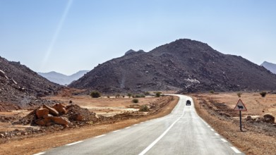 A car drives on a long road through a mountainous desert landscape under a blue sky, By car through