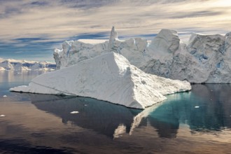 Large iceberg with striking reflection in calm water, surrounded by clouds and blue sky, icebergs