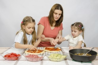 Mom helps younger daughter spread ketchup on a pizza, the eldest daughter, she is preparing a