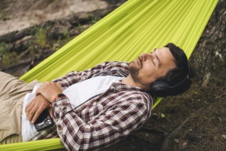 Man on bicycle trip at camping by lake is relaxing in green hammock while listening to music.