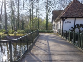 A wooden bridge leads to an old house by the water, surrounded by trees that cast shade on the