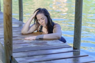 Beautiful woman bathing in a lake with her hands folded on a wooden pier looking at the sun