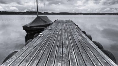 Wooden jetty and boat on a calm lake under a cloudy sky in black and white, Irsingener See, barrage