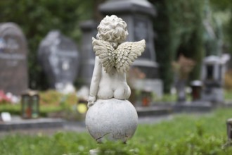 A little angel sits on a ball in a cemetery in front of graves in the blurred background