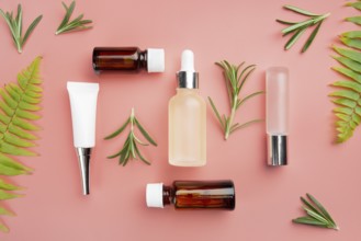 Glass and plastic cosmetic bottles on pink background with fern and rosemary leaves