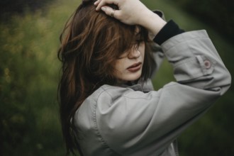 Portrait of a woman in a coat holding her hair, surrounded by nature with a mysterious vibe