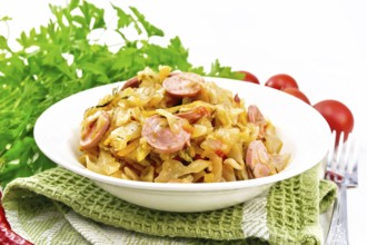 Cabbage stew with sausages in a white plate on a towel, tomatoes, parsley and a fork on the