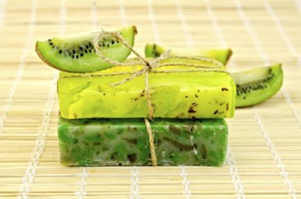 Two green homemade soap tied with twine, kiwi slices on a bamboo mat