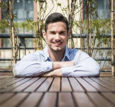Handsome young man resting crossed arms on wood surface while looking and smiling at camera