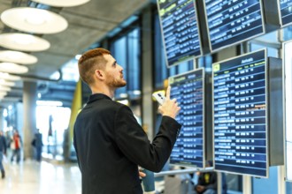 Businessman holding smartphone and pointing at timetable display, checking flight information in