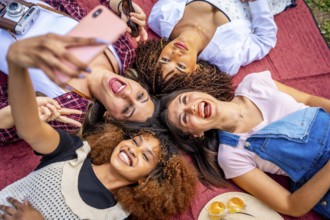 Four cheerful female friends are lying in a circle on a red blanket outdoors, taking a selfie with
