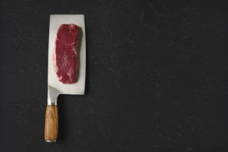 A piece of raw beef meat on a sharp cleaver with a wooden handle. The cleaver rests on a black