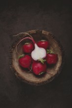 A collection of vibrant red radishes sits in a rustic wooden bowl. The contrasting dark background