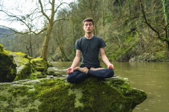 Yoga instructor meditating in lotus position on a mossy rock by a tranquil river, surrounded by