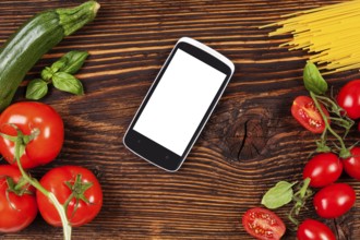 Fresh vegetables and pasta on wooden table with smartphone and recipe on screen. Groceries, recipe