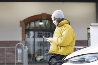 Woman with mobile phone in yellow jacket at charging station with electric car, environmentally