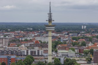 A tall television tower rises above a cityscape with numerous buildings, the sky is slightly